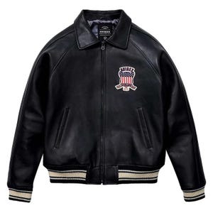 Peut inclure: Veste en cuir noire avec col, fermeture &eacute;clair et &eacute;cusson avec drapeau am&eacute;ricain et le mot "AVIREX". La veste a des poignets et un ourlet ray&eacute;s blancs et beiges.