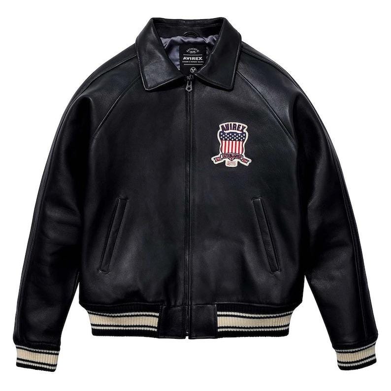 Peut inclure: Veste en cuir noire avec col, fermeture &eacute;clair et &eacute;cusson avec drapeau am&eacute;ricain et le mot "AVIREX". La veste a des poignets et un ourlet ray&eacute;s blancs et beiges.
