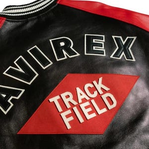 Op de afbeelding: Zwarte leren jas met rode accenten op de schouders en een zwart-witte gestreepte kraag. De jas heeft het woord "AVIREX" in grote, wit omrande zwarte letters en een rode ruit met "TRACK FIELD" in wit.