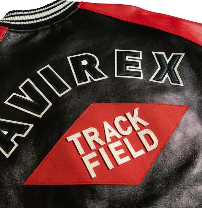 Op de afbeelding: Zwarte leren jas met rode accenten op de schouders en een zwart-witte gestreepte kraag. De jas heeft het woord "AVIREX" in grote, wit omrande zwarte letters en een rode ruit met "TRACK FIELD" in wit.