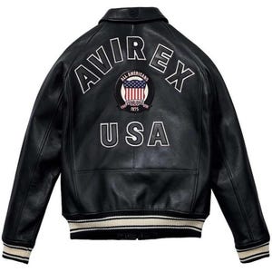 Peut inclure: Veste en cuir noire avec le mot "AVIREX" en arc de cercle sur les &eacute;paules et "USA" en dessous. Un &eacute;cusson circulaire avec le drapeau am&eacute;ricain est centr&eacute; entre les mots. La veste a un ourlet et des poignets c&ocirc;tel&eacute;s avec des rayures blanches.