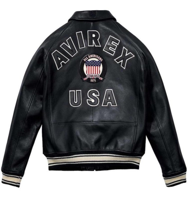 Peut inclure: Veste en cuir noire avec le mot "AVIREX" en arc de cercle sur les &eacute;paules et "USA" en dessous. Un &eacute;cusson circulaire avec le drapeau am&eacute;ricain est centr&eacute; entre les mots. La veste a un ourlet et des poignets c&ocirc;tel&eacute;s avec des rayures blanches.
