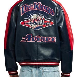 May include: A navy blue leather jacket with red and white accents. The back features the text "The Kings" above a crown graphic and the words "Gold Kickoff" and "Avirex". The jacket has a ribbed collar, cuffs, and hem.