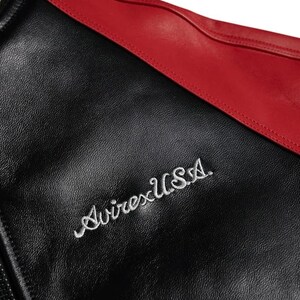 Op de afbeelding: Close-up van een zwarte leren jas met een rode accent. De jas heeft de geborduurde tekst "Avirex USA" in witte letters. Het leer heeft een getextureerde uitstraling.