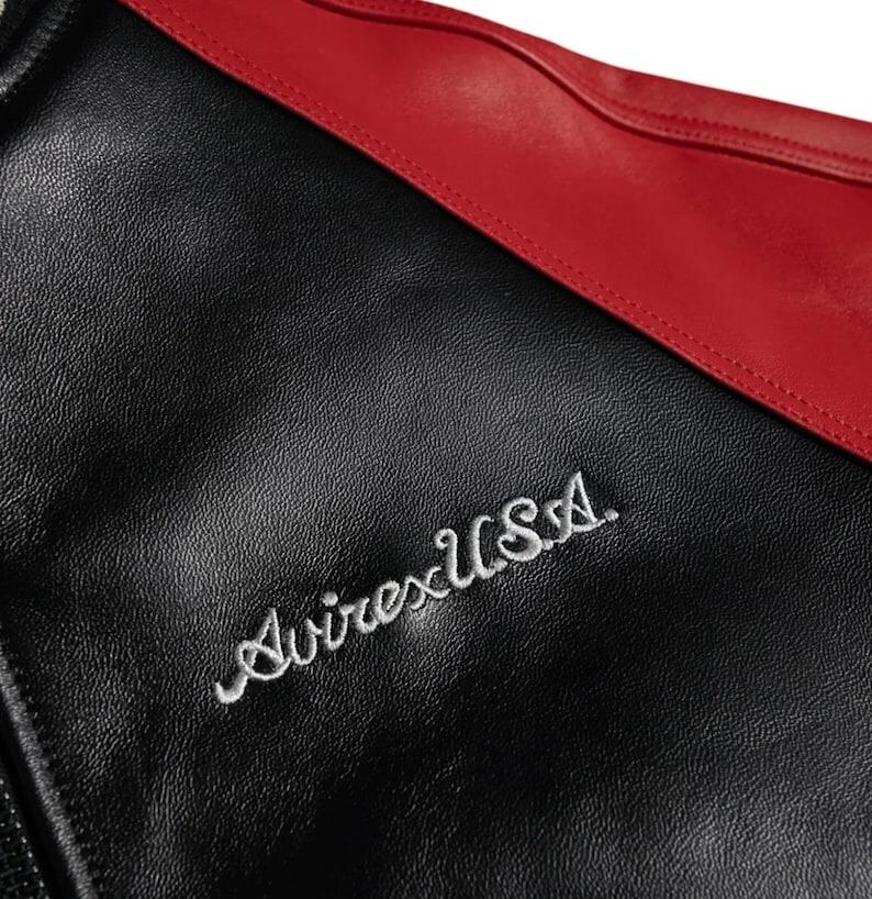 Op de afbeelding: Close-up van een zwarte leren jas met een rode accent. De jas heeft de geborduurde tekst "Avirex USA" in witte letters. Het leer heeft een getextureerde uitstraling.