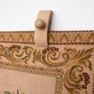 May include: Close-up of a tapestry with a decorative border and a wooden hanging mechanism. The tapestry features a beige background with floral and scroll patterns in shades of gold, green, and burgundy. The wooden hanger has a rounded knob.