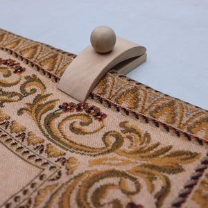 May include: A wooden table runner holder with a ball-shaped top, resting on a beige table runner. The runner features a detailed floral pattern in shades of gold, brown, and burgundy.