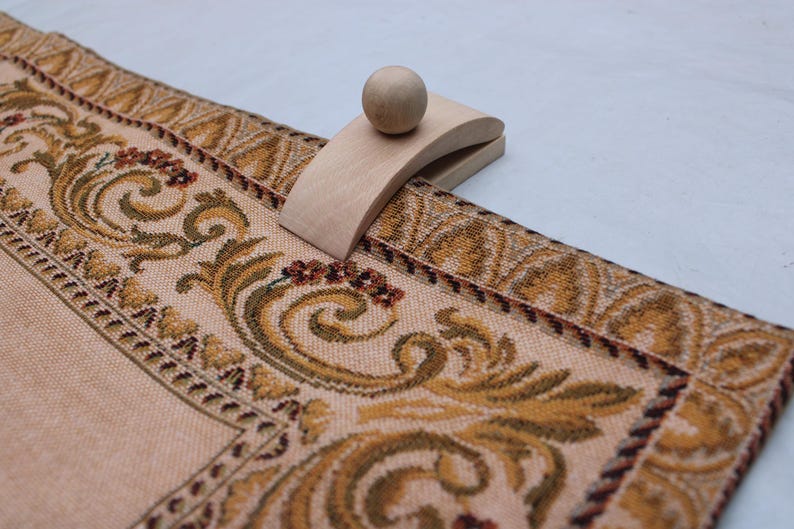 May include: A wooden table runner holder with a ball-shaped top, resting on a beige table runner. The runner features a detailed floral pattern in shades of gold, brown, and burgundy.