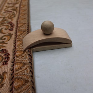 May include: A wooden rug beater with a rounded handle and a ball-shaped top. The beater is light-colored and sits next to a section of a patterned rug with brown, tan, and red floral designs.
