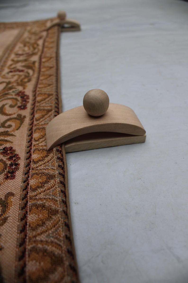 May include: A wooden rug beater with a rounded handle and a ball-shaped top. The beater is light-colored and sits next to a section of a patterned rug with brown, tan, and red floral designs.