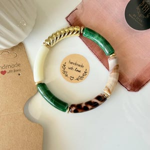 Può includere: Un braccialetto fatto a mano con una maglia a catena dorata, perline verdi, crema e stampa leopardata. Il braccialetto è esposto su una superficie bianca con un piccolo adesivo rotondo che dice "fatto a mano con amore."