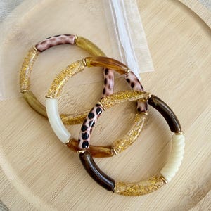 Peut inclure: Trois bracelets extensibles composés de perles cylindriques aux finitions variées, incluant des paillettes dorées, du blanc uni, du brun foncé, de l'ambre translucide et des motifs léopard, avec de petits anneaux intercalaires dorés.