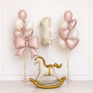 Puede incluir: Una composición festiva con globos y un caballito balancín. Los globos son rosas, blancos y transparentes, con forma de corazón y transparentes. Un caballito balancín dorado y blanco está en primer plano, ideal para un primer cumpleaños.
