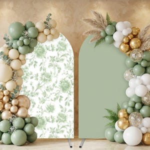 May include: Two arched backdrop stands, one with a floral pattern and the other solid green. Both are decorated with balloon garlands in shades of green, white, and gold, with accents of dried grasses and greenery. The stands are set against a neutral background.