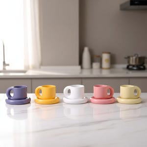 May include: A row of five colorful ceramic coffee cups and saucers. The cups are in shades of purple, yellow, white, pink, and pale yellow. The cups have a rounded handle and sit on matching round saucers. The background is a kitchen setting.
