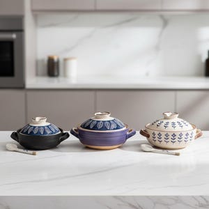 May include: Three ceramic cooking pots with lids and matching spoons. The pots are in black, purple, and cream colors, each with a unique leaf pattern on the lid. The pots have small handles and are set on a white countertop.