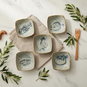 Puede incluir: Cinco cuencos cuadrados de cerámica con ilustraciones azules y blancas de criaturas marinas. Cada cuenco presenta un diseño diferente, incluyendo un pez, un calamar y un camarón. Los cuencos están dispuestos sobre un paño de color neutro con un tenedor de madera y ramas de olivo.