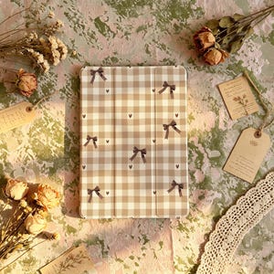 Può includere: Una custodia rettangolare per tablet con motivo a quadretti vichy beige e bianco, decorata con piccoli fiocchi marrone scuro e minuscole icone a forma di cuore distribuite sulla superficie. La custodia presenta un design ripiegabile.