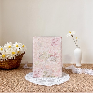 Op de afbeelding: Een roze tablet hoesje met bloemenpatroon en zwanenontwerp. De hoes heeft een delicaat patroon van roze bloemen en groene bladeren. Het rust op een wit kanten kleedje. Een mand met madeliefjes en een kleine vaas met bloemen staan op de achtergrond.