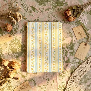 May include: A rectangular tablet case featuring a vertical striped pattern of yellow and light blue bands. The design includes small yellow floral motifs and delicate white dotted lines. The case has a tri-fold cover structure with visible seams.