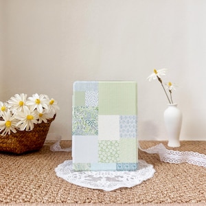 Może przedstawiać: Prostokątna okładka na notatnik w stylu patchwork w odcieniach zieleni, błękitu i bieli. Okładka wykonana jest z różnych wzorzystych tkanin, w tym pasków i wzorów kwiatowych. Notatnik spoczywa na białej koronkowej serwetce.