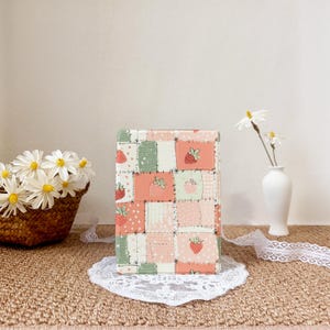 以下が含まれることがあります： 白い背景に、赤、ピンク、緑のイチゴが描かれたパッチワークデザインの長方形のブックカバー。白いレースのドイリーの上に置かれ、白い花のバスケットと背景に花瓶が置かれています。