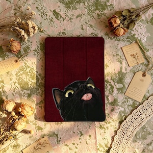 Puede incluir: Una funda para tableta de color burdeos intenso con una ilustración gráfica de un gato negro de ojos amarillos grandes y la lengua fuera. La funda tiene una superficie texturizada y un diseño de tres pliegues para proteger el dispositivo.