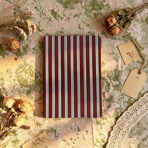 May include: A rectangular tablet case featuring a vertical striped pattern in deep burgundy, white, and light blue. The case has a smooth, flat surface with a folding seam visible on the left side, designed to protect a mobile electronic device.