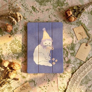 Puede incluir: Una funda rectangular de color morado para tableta con una ilustración central de un gato blanco que lleva un gorro de fiesta amarillo con lunares blancos y un cuello con volantes. El gato está sentado mirando al frente, con tres círculos.