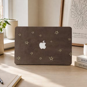 May include: A dark brown laptop skin featuring a scattered pattern of small, gold-colored star outlines. The skin is applied to the lid of a laptop, with the white Apple logo visible in the center, surrounded by the star design across the surface.