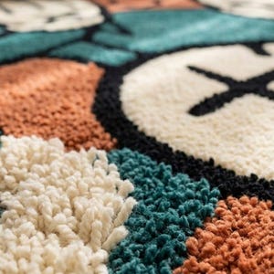May include: A tufted area rug featuring a geometric pattern of circular and block shapes in cream, teal, and burnt orange, outlined in thick black yarn. The surface has a high-pile, textured finish with visible loops of soft, dense yarn throughout.