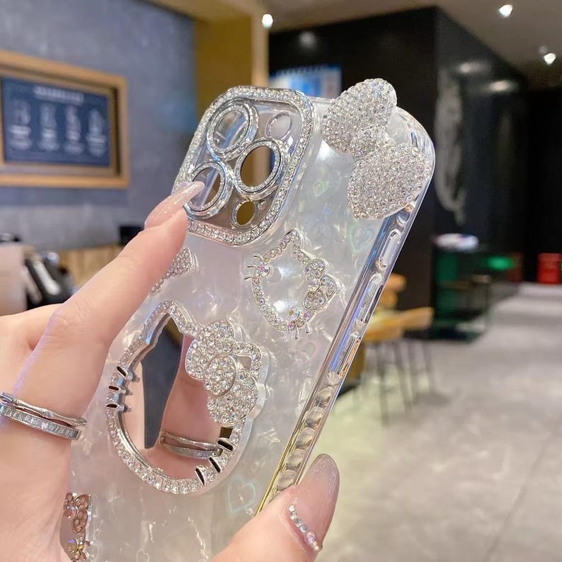 May include: Clear phone case with silver rhinestone embellishments. Features a bow, Hello Kitty designs, and a ring holder. The case is designed to fit a phone and has a pearlescent background.