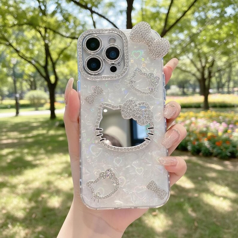 May include: A clear phone case with a white iridescent background, adorned with sparkling rhinestones. The case features a Hello Kitty design, bows, and a frame around the camera. The case is held in a hand.