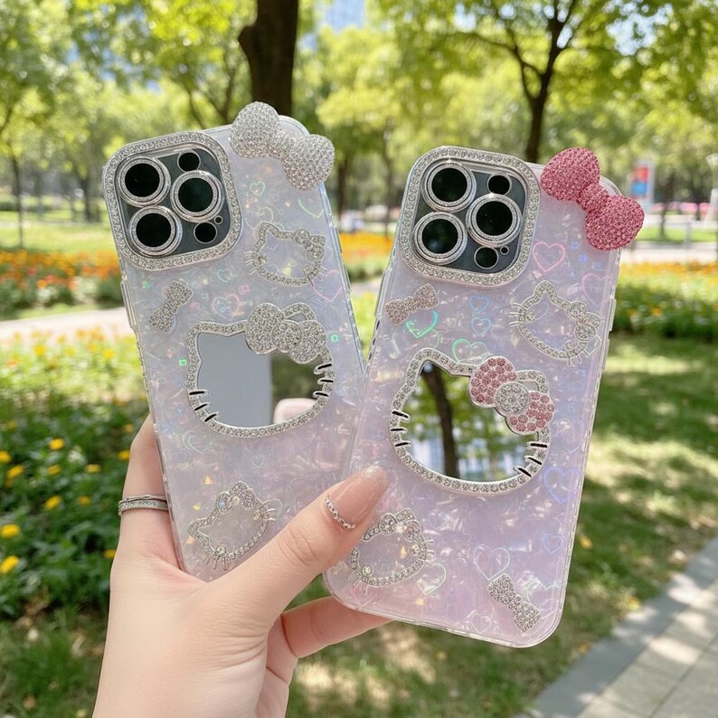 May include: Two phone cases with a Hello Kitty design. The cases are light purple with glitter and rhinestone accents. Each case has a bow at the top and a Hello Kitty face with a bow. The camera lens is surrounded by rhinestones.