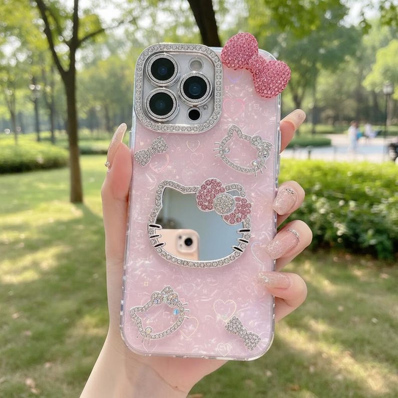 May include: Pink phone case with a Hello Kitty design. The case features a mirror, a sparkling bow, and rhinestone accents. The case is designed to fit a phone with multiple camera lenses.