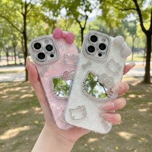 May include: Two phone cases, one pink and one white, are adorned with sparkling rhinestones and Hello Kitty designs. Each case features a bow and a mirror shaped like Hello Kitty's face. The cases are held up against a blurred green background.