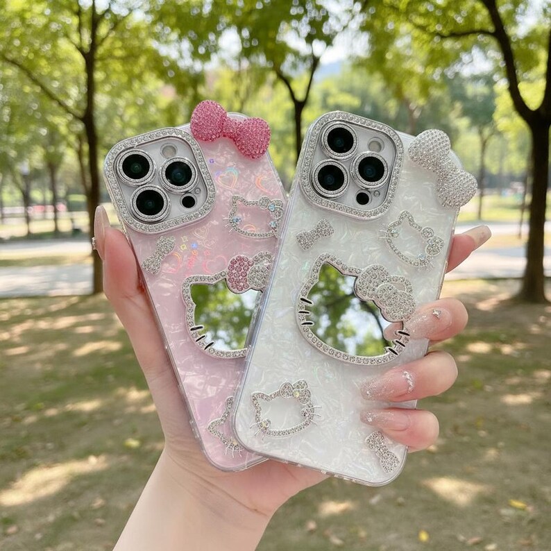 May include: Two phone cases, one pink and one white, are adorned with sparkling rhinestones and Hello Kitty designs. Each case features a bow and a mirror shaped like Hello Kitty's face. The cases are held up against a blurred green background.