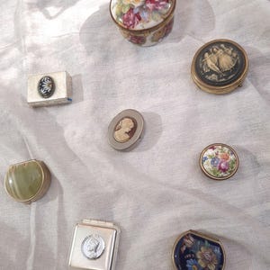 Puede incluir: Una colección de ocho pequeñas cajas decorativas y polveras con diversos diseños, incluyendo un tarro de porcelana con flores, una polvera con camafeo negro, una caja de piedra verde, una caja cuadrada plateada con un retrato y piezas de esmalte floral.