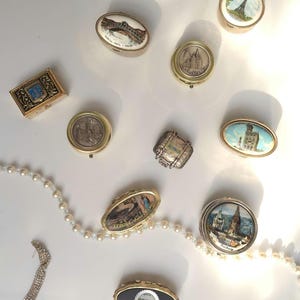Può includere: Una collezione di piccole scatole portapillole e contenitori decorativi in metallo con vari monumenti e scene urbane, tra cui la Torre Eiffel e il Ponte di Rialto, accompagnati da una collana di perle bianche e una spilla con strass.