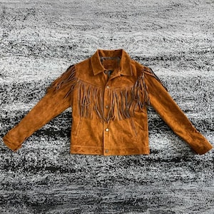 Könnte beinhalten: Braune Wildlederjacke mit langen Ärmeln und Knopfleiste. Die Jacke hat dekorative Fransen über der Brust und den Schultern. Die Jacke liegt flach auf einer strukturierten Oberfläche.