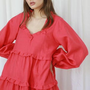 May include: Coral-colored dress with long, voluminous sleeves and a tiered, ruffled skirt. The dress features a V-neckline with delicate ruffle detailing and a cinched waist. The fabric appears to be a lightweight linen or cotton.