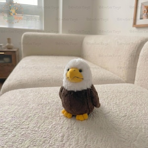 Puede incluir: Un peluche de águila calva con cabeza blanca, cuerpo marrón y pico y patas amarillas. El peluche está sobre una superficie blanca texturizada. Es un artículo para niños.