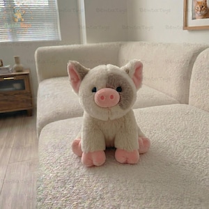 May include: A plush pig toy with cream-colored fur, a soft pink snout, and pink hooves. The stuffed animal features a prominent gray patch over its left eye and ear, with dark, round eyes and upright, pointed ears, sitting in a seated position.