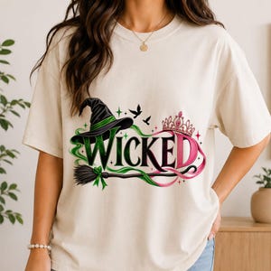 Op de afbeelding: Een crèmekleurig T-shirt met korte mouwen en het woord WICKED in grote groene en roze letters. Het ontwerp bevat een zwarte heksenhoed, een bezemsteel, een roze tiara en kleine zwarte vogelsilhouetten, geaccentueerd met groene krullen.