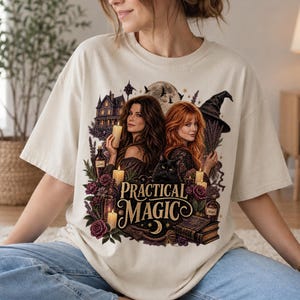 May include: A cream-colored t-shirt featuring a graphic print of two women with long hair, a black cat, a witch hat, a house, candles, flowers, and books. The text "PRACTICAL MAGIC" is printed in a decorative font in the center of the design.