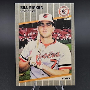 May include: A 1989 Fleer baseball card featuring Bill Ripken of the Baltimore Orioles. The player wears a white jersey with orange and black trim, holding a bat with the text 'Fuck Face' written on the knob. The card includes the team logo.