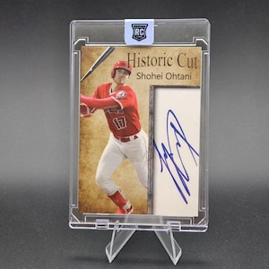 May include: A Shohei Ohtani Historic Cut rookie baseball card encased in a clear plastic holder. The card features a photograph of the player in a red jersey with the number 17, a blue ink autograph on a white background, and a Rookie RC sticker.