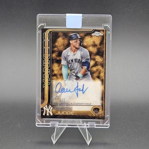 May include: A Topps Chrome baseball card featuring Aaron Judge in a New York Yankees uniform, encased in a clear plastic holder. The card displays a blue ink autograph, the text YANKEES, and a serial number 07/10 on a gold patterned background.