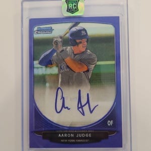 May include: A Bowman Chrome baseball trading card featuring Aaron Judge of the New York Yankees in a batting stance. The card has a purple border, a blue ink autograph, and a holographic Rookie RC sticker at the top, all encased in a clear plastic holder.