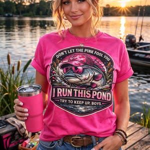 May include: A pink t-shirt with the text "DON'T LET THE PINK FOOL YOU" and "I RUN THIS POND TRY TO KEEP UP, BOYS." features a graphic of a catfish wearing sunglasses and a hat. The person is wearing blue jeans and holding a pink tumbler.
